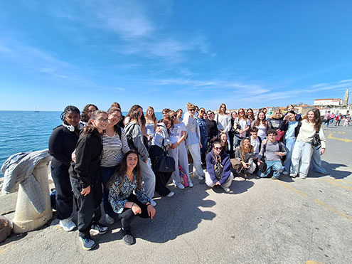 Velika skupina nasmejanih dijakov pozira na obalni ploščadi v Piranu. Nebo je popolnoma jasno. V ozadju se v daljavi vidi veduta Pirana s cerkvenim zvonikom.