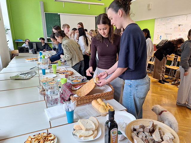 Dijaki se družijo ob dolgi mizi, polni hrane, v učilnici z živahno zelenimi stenami. Na mizah so tipične slovenske jedi: različne vrste kruha, prigrizki, piškoti in steklenica oljčnega olja. 