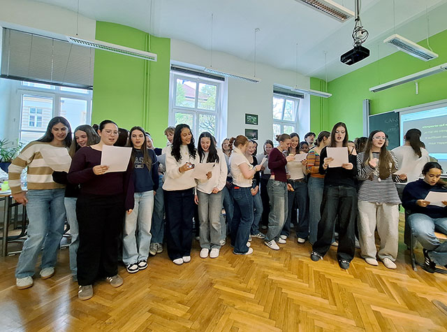 V isti zeleni učilnici velika skupina dijakov stoji skupaj in prepeva pesem Čez Šuštarski most - nekaj kitic v francoščini, refren v slovenščini. V rokah držijo liste z besedilom. Nekateri se smehljajo, drugi so osredotočeni na besedilo. 