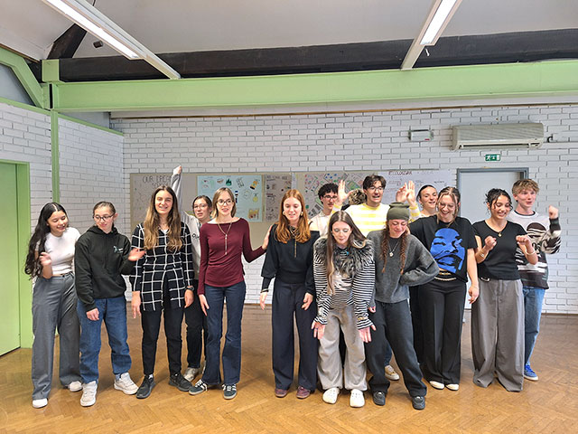 Skupina mladih stoji v učilnici z belo opečnato steno in rjavim parketom. Nekateri krilijo z rokami ali se premikajo, kot da bi bili sredi igre ali improvizacijskega nastopa. Vsi so videti dobre volje in polni energije.