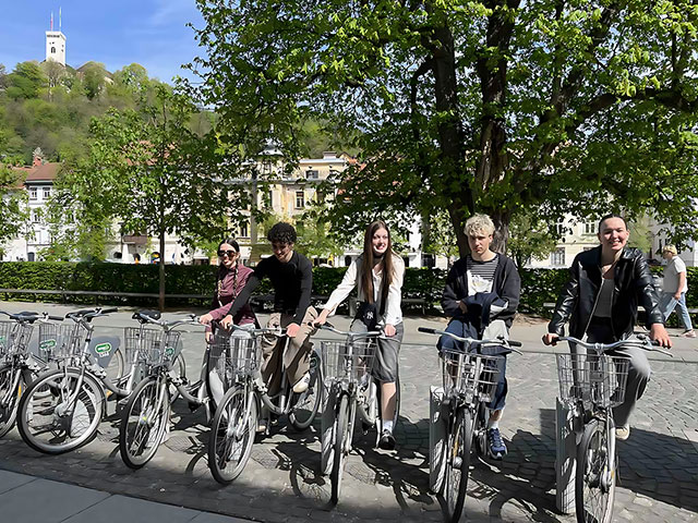 Pet dijakov sedi na mestnih kolesih sistema BicikeLJ na trgu v središču Ljubljane. V ozadju se nad mestom na zelenem hribu vidi Ljubljanski grad pod modrim nebom brez oblačkov.