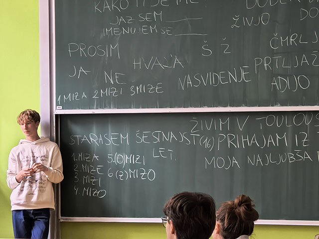 Dijak stoji ob šolski tabli, na kateri so s kredo zapisane osnovne slovenske besede in fraze, kot so "Jaz sem...", "Hvala", "Nasvidenje" in "Prosim". Dijako vodi delavnico slovenščine za francoske dijake. V spodnjem delu slike sta vidni glavi dveh dijakov, ki pozorno spremljata razlago.