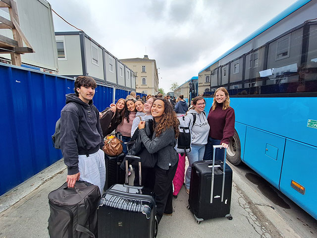 Nasmejana skupina mladih stoji na ozki poti med modrim potovalnim avtobusom in modro gradbiščno ograjo. Vsi držijo ali stojijo ob svojih kovčkih. Nekateri se objemajo in zrejo v objektiv, polni vtisov, pripravljeni na pot domov.