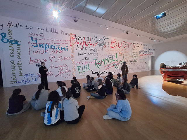 Skupina dijakov sedi na tleh prostorne galerije. Pred njimi je ogromna bela stena, prekrita s pisanimi napisi v različnih jezikih in političnimi gesli (npr. "Baghdad", "Ukraine", "America"). Ob strani stoji vodička, ki skupini razlaga razstavljeno umetnino, na desni pa je videti del instalacije s prevrnjenim rdečim avtomobilom.