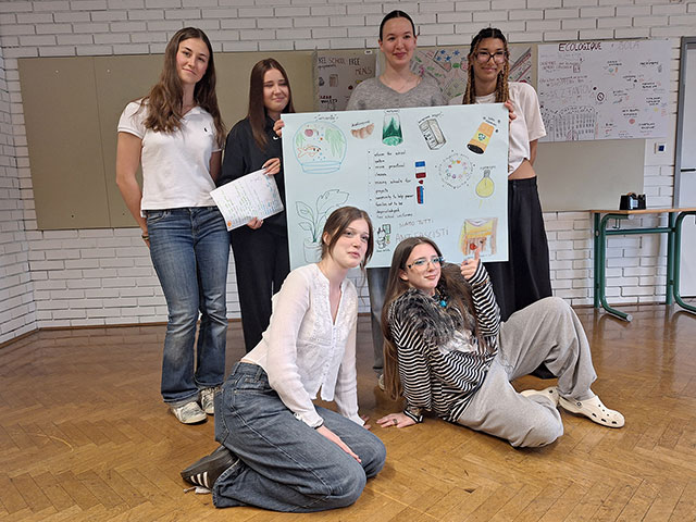 Šest dijakinj v učilnici ponosno drži velik ročno porisan plakat z naslovom "Our Dream School". Na plakatu so ilustracije akvarija, rastlin in žarnice ter seznam idej v angleščini, kot so "more practical classes" in "free school uniforms".