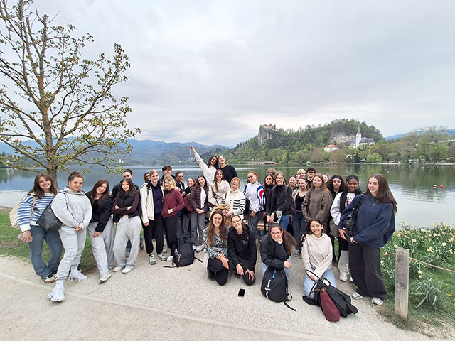 Velika skupina dijakov pozira na obali Blejskega jezera. V ozadju se na strmi pečini dviga Blejski grad, ob njem pa je vidna cerkev sv. Martina. Nebo je oblačno, ob robu poti pa cvetijo narcise.