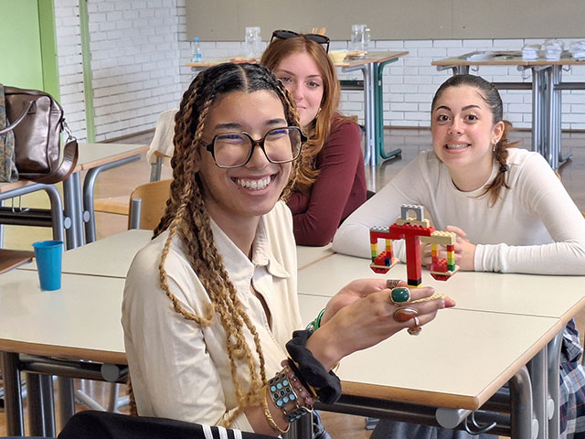 Tri dijakinje sedijo za šolsko mizo. Dijakinja v ospredju se široko smeji in v rokah drži majhno tehtnico, ki so jo sestavili iz pisanih lego kock. Vzdušje v učilnici je sproščeno in ustvarjalno.