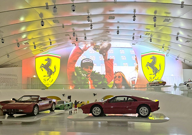 V ospredju so na belih ploščadih razstavljeni klasični športni avtomobili Ferrari. V ozadju je veliko ukrivljeno stensko platno, na katerem se predvaja projekcija – videti je dirkača s pokalom v rokah, obdanega z lovorovim vencem, na obeh straneh pa sta prepoznavna rumena logotipa Ferrarija s vzpenjajočim se konjem. Prostor ima moderen dizajn z obokanim stropom in številnimi reflektorji, ki osvetljujejo eksponate.