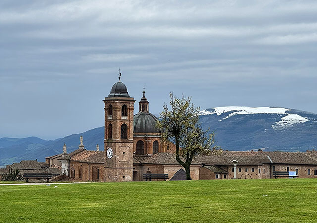 Fotografija prikazuje razgled na zgodovinsko središče mesta Urbino. V ospredju je zelena trata, v ozadju pa se dvigajo stolnica s kupolo in zvonikom ter zasneženi vrhovi Apeninov.