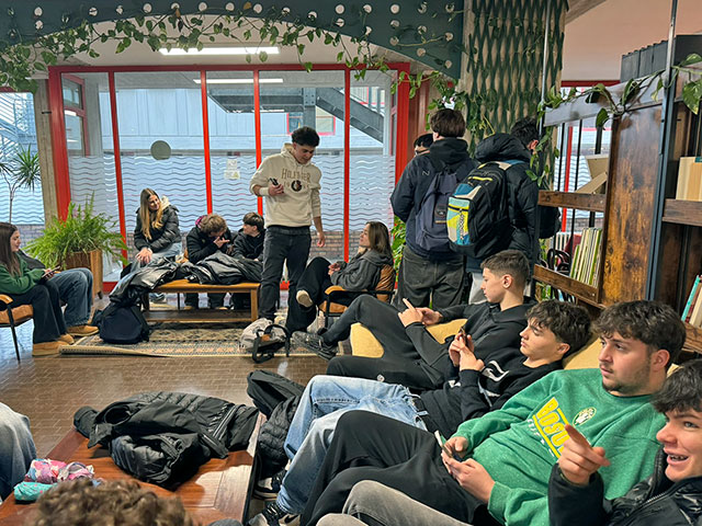 Slovenski in italijanski dijaki v sproščenem vzdušju večinoma sedijo v šolski avli. Pogovarjajo se ali pa brskajo po telefonih.