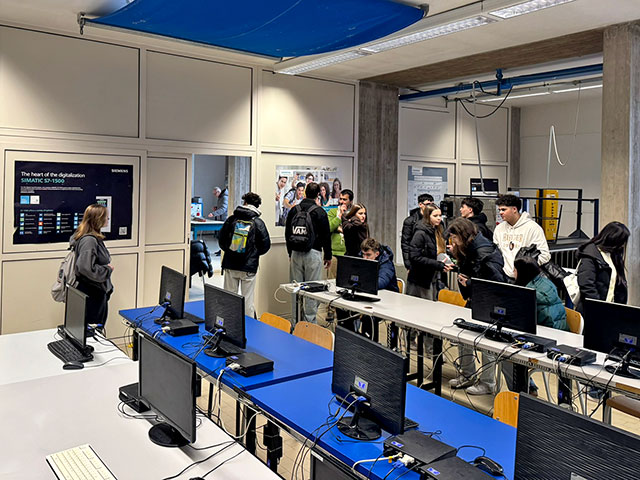 Notranjost računalniške učilnice z več vrstami miz, na katerih so nameščeni računalniški monitorji. V prostoru je skupina dijakov, ki si ogledujejo učilnico.