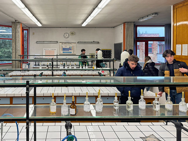 Fotografija prikazuje šolski kemijski laboratorij. V ospredju je dolga laboratorijska miza s steklenimi policami, na katerih so v vrsti razporejene stekleničke s kemikalijami, opremljene s kapalkami. V ozadju so vidne druge laboratorijske mize in stena s kemijskimi pripomočki. V prostoru je šest dijakov, ki si ogledujejo laboratorij.