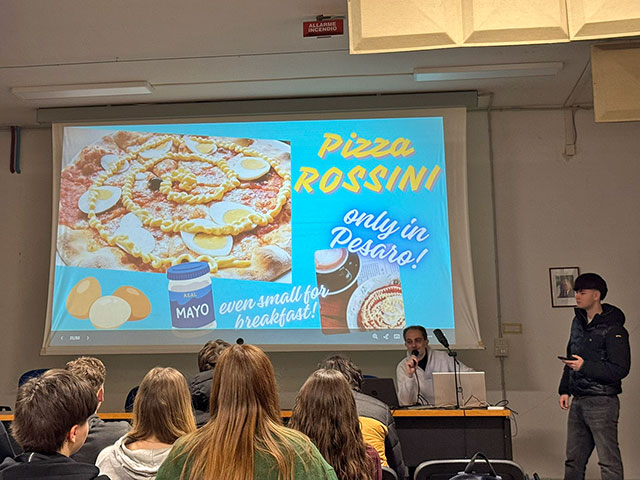 Prizor v predavalnici, kjer poteka predstavitev. Na velikem projekcijskem platnu je slika pice z jajci in napisom "Pizza ROSSINI, only in Pesaro!". Za mizo sedi profesor z mikrofonom, ob njem pa dijak. Ostali dijaki poslušajo predavanje.