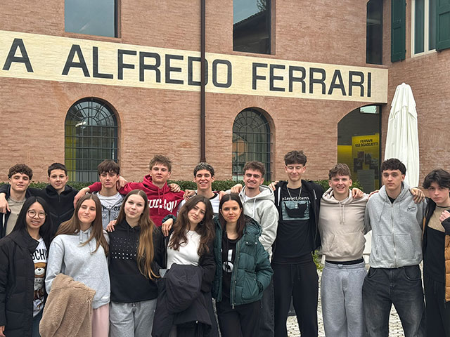 Skupinska fotografija 15 dijakov, ki pozirajo pred opečnato stavbo Ferrarijevega muzeja. Stojijo v dveh vrstah, so sproščeni in nasmejani.