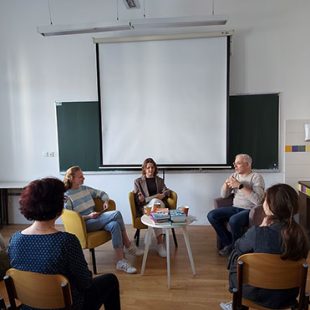 Trije sogovorniki, to sta dve dijakinji in Žiga Valetič, sedijo v naslanjačih pred belim projekcijskim platnom v učilnici. Žiga Valetič razlaga, odgovarja na vprašanja. Na nizki mizici med njimi je zložen kup knjig. V ospredju so vidni dijaki, ki dogodek spremljajo s stolov.