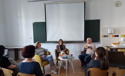 Trije sogovorniki, to sta dve dijakinji in Žiga Valetič, sedijo v naslanjačih pred belim projekcijskim platnom v učilnici. Žiga Valetič razlaga, odgovarja na vprašanja. Na nizki mizici med njimi je zložen kup knjig. V ospredju so vidni dijaki, ki dogodek spremljajo s stolov.