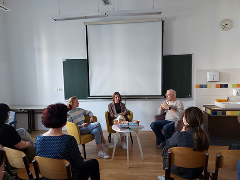 Trije sogovorniki, to sta dve dijakinji in Žiga Valetič, sedijo v naslanjačih pred belim projekcijskim platnom v učilnici. Žiga Valetič razlaga, odgovarja na vprašanja. Na nizki mizici med njimi je zložen kup knjig. V ospredju so vidni dijaki, ki dogodek spremljajo s stolov.