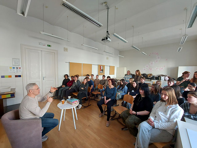 Žiga Valetič sedi na naslovnjaču v prednjem delu učilnice. Na okrogli mizi pred njim je kup pisanih knjig. Rzlaga in gestikulira z rokami. Njemu nasproti sedijo dijaki in profesorica ter poslušajo njegovo razlago.
