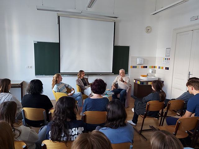 Širok posnetek učilnice med pogovorom z Žigo Valetičem, ki sedi v naslanjaču in med govorom gestikulira. Na njegovi desni sedita dve dijakinji, vi vodita pogovor znjim. Pred njimi na stolih sedi večje število dijakov.