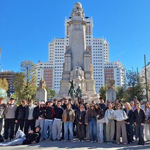 Velika skupina približno 30 dijakov in učiteljev pozira za skupinski portret na sončen dan. Oblečeni so v jesenska oblačila (puloverji, kavbojke, lahke jakne). Eden izmed dijakov v ospredju na levi humorno leži na tleh. Fotografija je posneta na odprtem trgu Plaza de España v Madridu. Nebo je popolnoma jasno, kar ustvarja živahno in pozitivno popotniško vzdušje. Na levi in desni strani so vidna mestna drevesa in moderni nebotičniki, ki obdajajo trg.