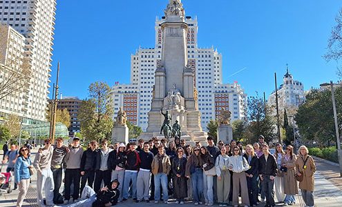 Velika skupina približno 30 dijakov in učiteljev pozira za skupinski portret na sončen dan. Oblečeni so v jesenska oblačila (puloverji, kavbojke, lahke jakne). Eden izmed dijakov v ospredju na levi humorno leži na tleh. Fotografija je posneta na odprtem trgu Plaza de España v Madridu. Nebo je popolnoma jasno, kar ustvarja živahno in pozitivno popotniško vzdušje. Na levi in desni strani so vidna mestna drevesa in moderni nebotičniki, ki obdajajo trg.