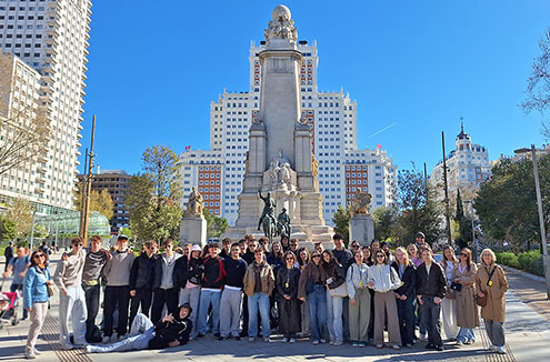 Velika skupina približno 30 dijakov in učiteljev pozira za skupinski portret na sončen dan. Oblečeni so v jesenska oblačila (puloverji, kavbojke, lahke jakne). Eden izmed dijakov v ospredju na levi humorno leži na tleh. Fotografija je posneta na odprtem trgu Plaza de España v Madridu. Nebo je popolnoma jasno, kar ustvarja živahno in pozitivno popotniško vzdušje. Na levi in desni strani so vidna mestna drevesa in moderni nebotičniki, ki obdajajo trg.