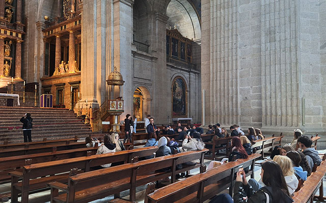 Fotografija prikazuje notranjost monumentalne bazilike El Escorial. V ospredju in sredini so dijaki, ki sedijo v dolgih, temnih lesenih klopeh. Nekateri pozorno poslušajo, drugi fotografirajo s svojimi telefoni. Pred njimi stoji vodič, ki jim nekaj razlaga. Prostor zaznamujejo masivni sivi kamniti stebri in visoki oboki. Na levi strani je viden velik zlat oltar s kipi in stebriči, poleg njega pa se dviga visoka, bogato okrašena zlata prižnica.
V ozadju na stenah visijo velike nabožne slike v masivnih okvirjih.