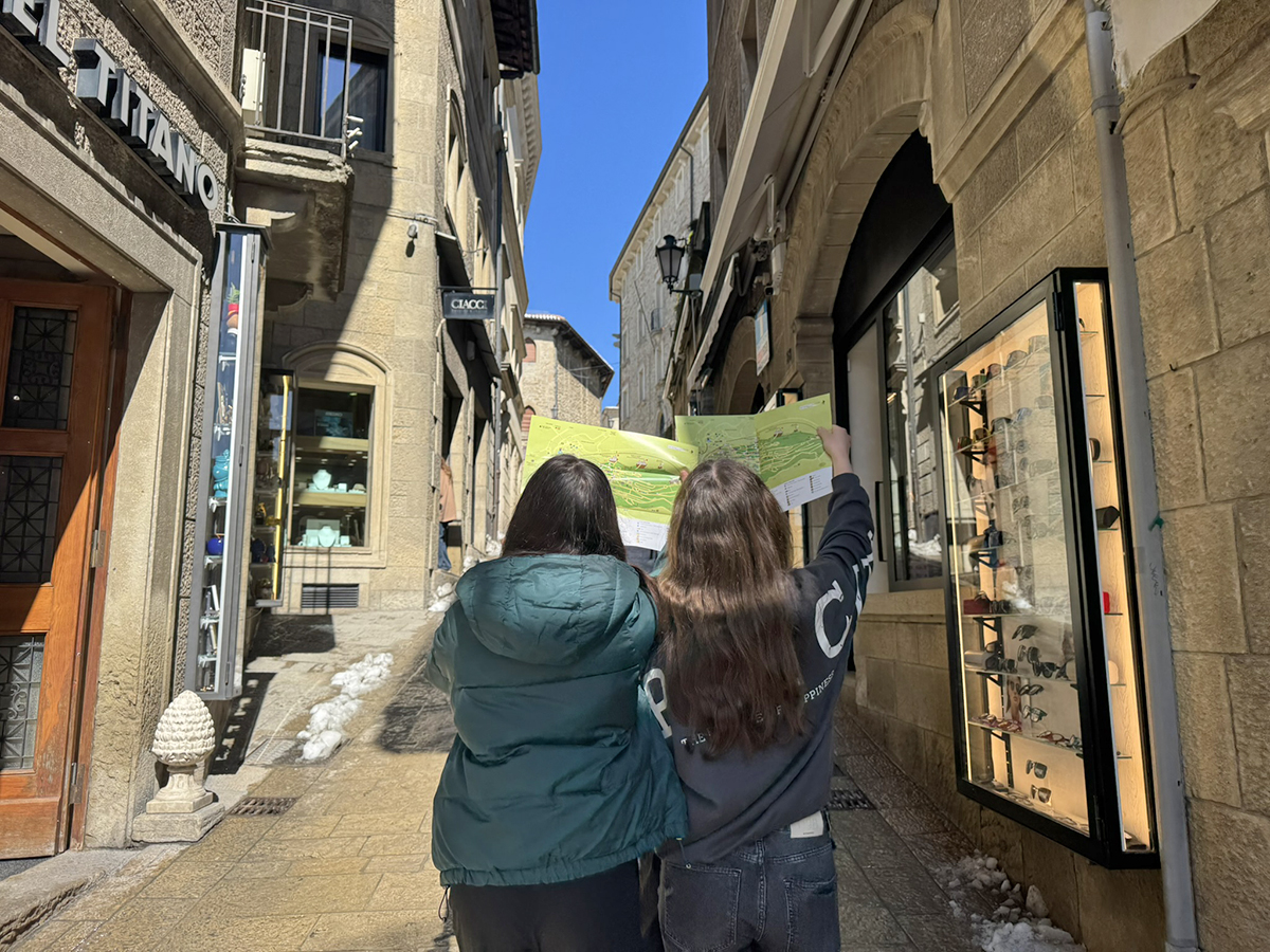 Dve dijakinji stojita na strmi, tlakovani ulici med starimi kamnitimi stavbami. Vidimo ju v hrbet, pred seboj držita razprt turistični zemljevid. Ulica je osvetljena s soncem, ob straneh pa sta vidni izložbi trgovin.