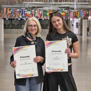 V prostornem zaprtem hodniku stojita nasmejani mentorica in nagrajena dijakinja. V rokah ponosno držita uradni priznanji. Oblečeni sta v temnejša oblačila. V ozadju nad njima visi dolga vrsta mednarodnih zastav, prostor pa dopolnjujejo beli stebri in svetla tla.