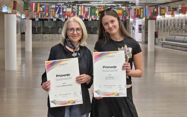 V prostornem zaprtem hodniku stojita nasmejani mentorica in nagrajena dijakinja. V rokah ponosno držita uradni priznanji. Oblečeni sta v temnejša oblačila. V ozadju nad njima visi dolga vrsta mednarodnih zastav, prostor pa dopolnjujejo beli stebri in svetla tla.