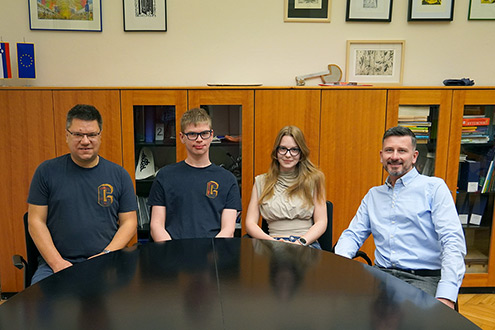 Skupinski portret štirih oseb – dveh profesorjev mentorjev in dijaka ter dijakinje, ki sedita med profesorjema. Vsi sedijo za temno konferenčno mizo, v ozadju so lesene omare s knjigami, uokvirjene slike na steni ter zastavi Slovenije in Evropske unije.