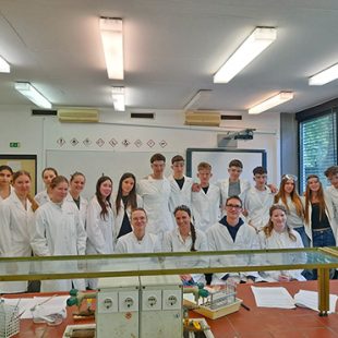 Skupinski portret dijakov in mentoric v kemijskem laboratoriju na Pedagoški fakulteti v Ljubljani. Vsi nosijo bele zaščitne halje, nekateri imajo na glavi tudi zaščitna očala. Nasmejani stojijo in sedijo za laboratorijsko mizo, v ozadju sta bela tabla s piktogrami za nevarnosti in projekcijsko platno.