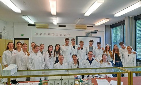 Skupinski portret dijakov in mentoric v kemijskem laboratoriju na Pedagoški fakulteti v Ljubljani. Vsi nosijo bele zaščitne halje, nekateri imajo na glavi tudi zaščitna očala. Nasmejani stojijo in sedijo za laboratorijsko mizo, v ozadju sta bela tabla s piktogrami za nevarnosti in projekcijsko platno.