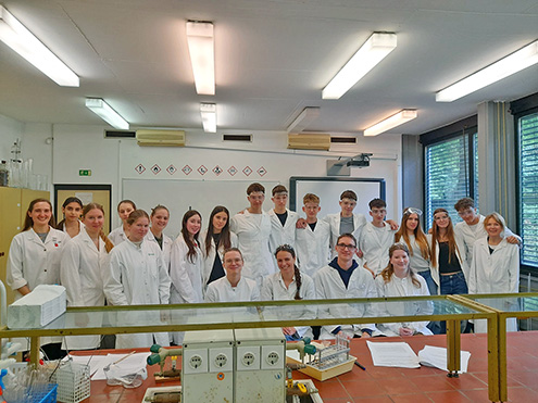 Skupinski portret dijakov in mentoric v kemijskem laboratoriju na Pedagoški fakulteti v Ljubljani. Vsi nosijo bele zaščitne halje, nekateri imajo na glavi tudi zaščitna očala. Nasmejani stojijo in sedijo za laboratorijsko mizo, v ozadju sta bela tabla s piktogrami za nevarnosti in projekcijsko platno.