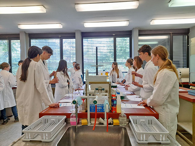 Dijaki v belih haljah med delom pri laboratorijskih mizah. Razdeljeni so v manjše skupine, prebirajo delovne liste in si ogledujejo laboratorijske pripomočke. V ospredju je vidno veliko pomivalno korito s stojali za sušenje posode, v ozadju pa so velika okna z žaluzijami.