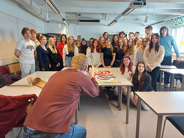 Fotograf v ospredju slika veliko skupino mladih, ki pozirajo v učilnici ob mizi z barvito umetnino, ki so jo ustvarili skupaj.