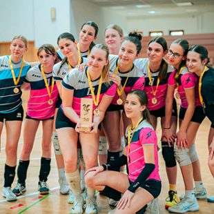 Skupinski posnetek nasmejane dekliške odbojkarske ekipe Gimnazije Ledina, ki v športni dvorani proslavlja uspeh z medaljami okoli vratu in pokalom v rokah.