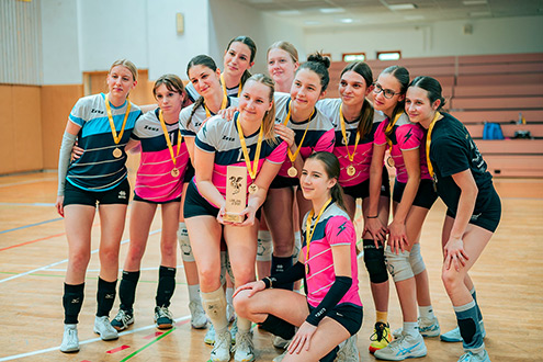 Skupinski posnetek nasmejane dekliške odbojkarske ekipe Gimnazije Ledina, ki v športni dvorani proslavlja uspeh z medaljami okoli vratu in pokalom v rokah.