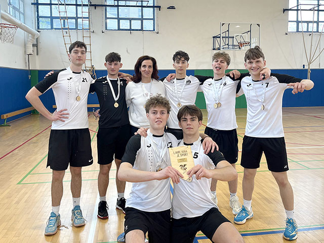 Zmagoslavna fotografija fantovske odbojkarske ekipe Gimnazije Ledina s trenerko, kjer igralci ponosno kažejo osvojene medalje in priznanje za dosežen športni uspeh.