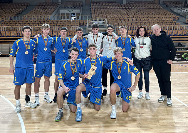 Zmagovalna rokometna ekipa Gimnazije Ledina z bronastimi medaljami in pokalom pozira na igrišču velike dvorane v spremstvu trenerke.