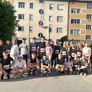 Skupinska fotografija približno tridesetih mladih tekačev, ki so se postavili v dve vrsti – stoječo in klečečo. Večina jih je oblečena v športna oblačila z vidnimi štartnimi številkami. V ozadju je vidna stanovanjska stavba z neobičajnimi okroglimi okni na levi in običajnimi pravokotnimi okni na desni strani.