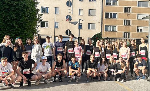 Skupinska fotografija približno tridesetih mladih tekačev, ki so se postavili v dve vrsti – stoječo in klečečo. Večina jih je oblečena v športna oblačila z vidnimi štartnimi številkami. V ozadju je vidna stanovanjska stavba z neobičajnimi okroglimi okni na levi in običajnimi pravokotnimi okni na desni strani.