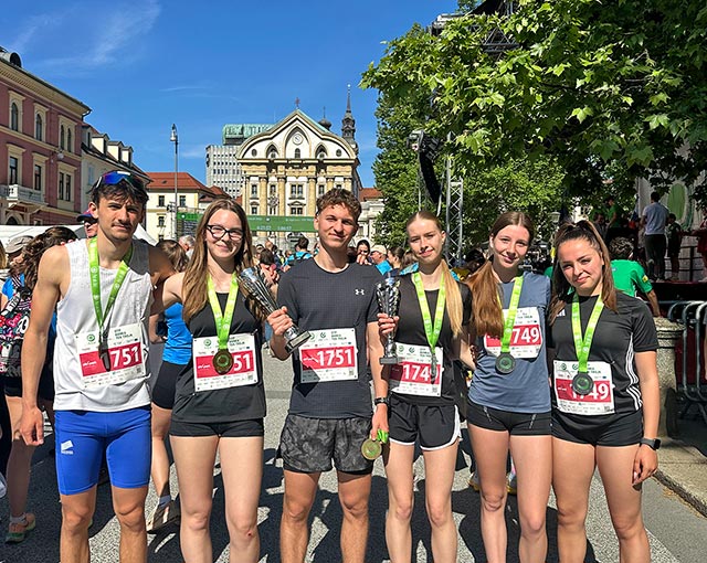 Šest mladih tekačev, trije fantje in tri dekleta, pozira na mestnem trgu. Vsi imajo okoli vratu medalje, dva med njimi pa v rokah držita pokale. V ozadju se vidi cerkev, druge starejše zgradbe in zelena drevesa.