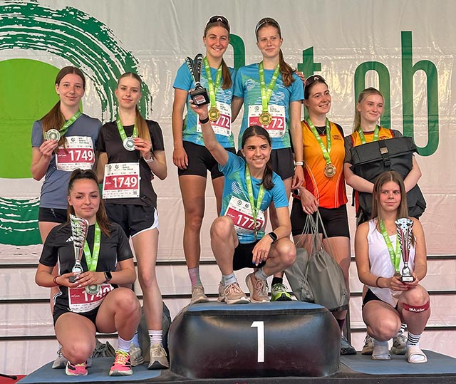 Podelitev medalj in pokalov skupini devetih mladih tekačic, ki so se razvrstile na treh stopnjah zmagovalnih stopničk. Tri dekleta čepijo v ospredju, šest pa jih stoji za njimi. Vse imajo medalje okoli vratu, tri izmed njih pa v rokah držijo srebrne pokale.