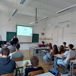 Fotografija prikazuje šolsko učilnico med predavanjem. Predavatelj stoji ob projekcijskem platnu s predstavitvijo, pred njim pa v klopeh sedi skupina dijakov, ki spremlja razlago.
