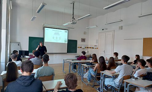 Fotografija prikazuje šolsko učilnico med predavanjem. Predavatelj stoji ob projekcijskem platnu s predstavitvijo, pred njim pa v klopeh sedi skupina dijakov, ki spremlja razlago.