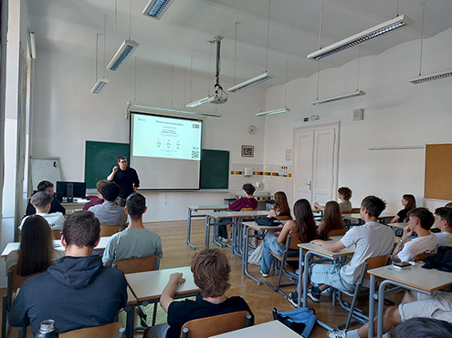 Fotografija prikazuje šolsko učilnico med predavanjem. Predavatelj stoji ob projekcijskem platnu s predstavitvijo, pred njim pa v klopeh sedi skupina dijakov, ki spremlja razlago.