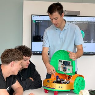 Mentor dvema dijakoma podrobno kaže elektronsko vezje in notranjost odprtega oranžno-zelenega robotskega vozila na mizi.