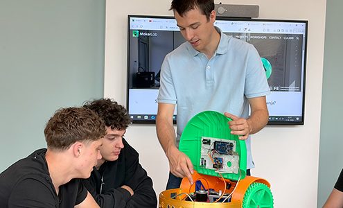 Mentor dvema dijakoma podrobno kaže elektronsko vezje in notranjost odprtega oranžno-zelenega robotskega vozila na mizi.