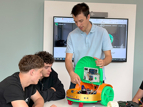 Mentor dvema dijakoma podrobno kaže elektronsko vezje in notranjost odprtega oranžno-zelenega robotskega vozila na mizi.